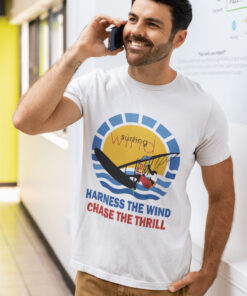 Man wearing a white windsurfing t-shirt with a colorful design of a windsurfer gliding over waves against a sun backdrop, paired with the phrases 'Harness the Wind' and 'Chase the Thrill