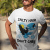 A smiling man with sunglasses and a beard wearing a white premium kite surfing t-shirt on a tropical beach. The t-shirt features a kitesurfing silhouette with the phrase "Salty Hair, Don’t Care," along with blue waves, a yellow sun, and swirl elements, capturing the adventurous ocean lifestyle.