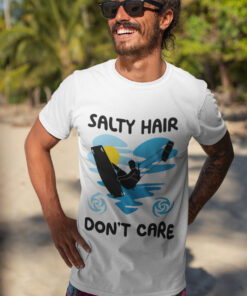 A smiling man with sunglasses and a beard wearing a white premium kite surfing t-shirt on a tropical beach. The t-shirt features a kitesurfing silhouette with the phrase "Salty Hair, Don’t Care," along with blue waves, a yellow sun, and swirl elements, capturing the adventurous ocean lifestyle.