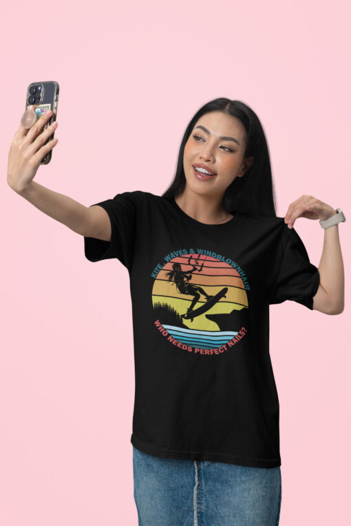 Waves & Windblown Hair – Epic Kite Surfing T-Shirt featuring a bold silhouette of a female kitesurfer soaring over a scenic sunset landscape with a retro-style striped background. The design includes the phrases "KITE, WAVES & WINDBLOWN HAIR" in blue and "WHO NEEDS PERFECT NAILS?" in red, adding a fun, adventurous touch.