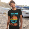 Epic Kite Surfing T-Shirt – Ocean Playground Adventure | A young man with a smile stands on a sandy beach near boats, wearing a black kite surfing t-shirt with a vibrant retro sunset design featuring a kitesurfer silhouette. The text "OCEAN PLAYGROUND KITE INCLUDED" is printed in bold colors, capturing the adventurous spirit of kite surfing