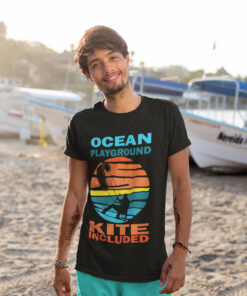 Epic Kite Surfing T-Shirt – Ocean Playground Adventure | A young man with a smile stands on a sandy beach near boats, wearing a black kite surfing t-shirt with a vibrant retro sunset design featuring a kitesurfer silhouette. The text 