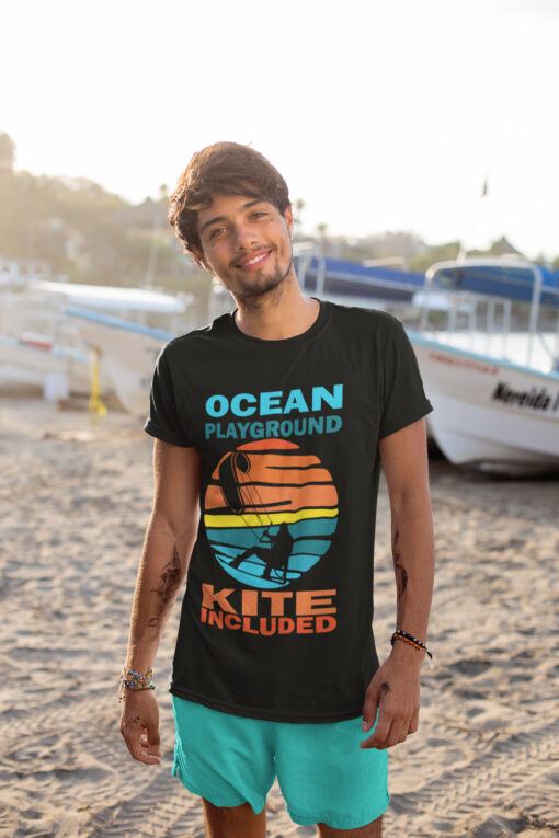 Epic Kite Surfing T-Shirt – Ocean Playground Adventure | A young man with a smile stands on a sandy beach near boats, wearing a black kite surfing t-shirt with a vibrant retro sunset design featuring a kitesurfer silhouette. The text "OCEAN PLAYGROUND KITE INCLUDED" is printed in bold colors, capturing the adventurous spirit of kite surfing