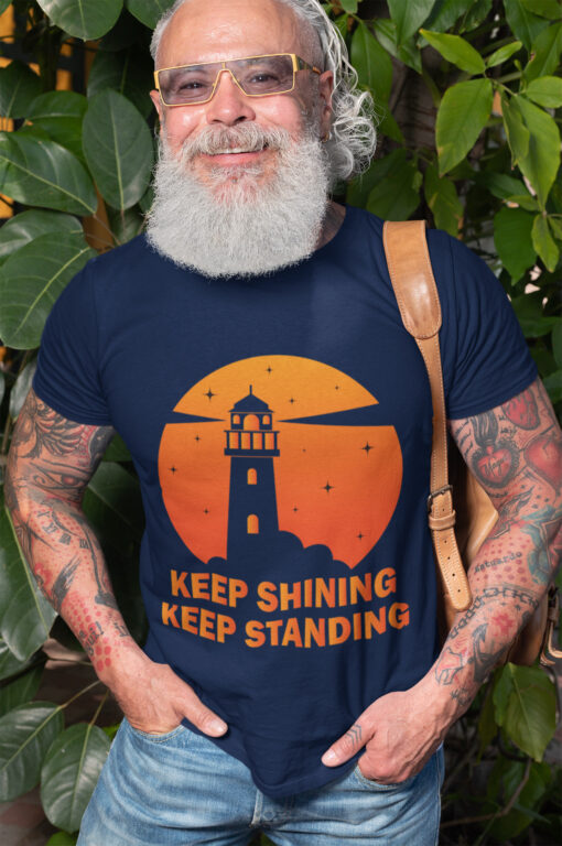 A senior man with a white beard, sunglasses, and a trendy style wears a navy blue "Keep Shining, Keep Standing" nautical t-shirt. The shirt features a bold lighthouse silhouette against an orange sunset with scattered stars, symbolizing resilience and strength. He stands confidently with a leather backpack strap over his shoulder, surrounded by lush green foliage.