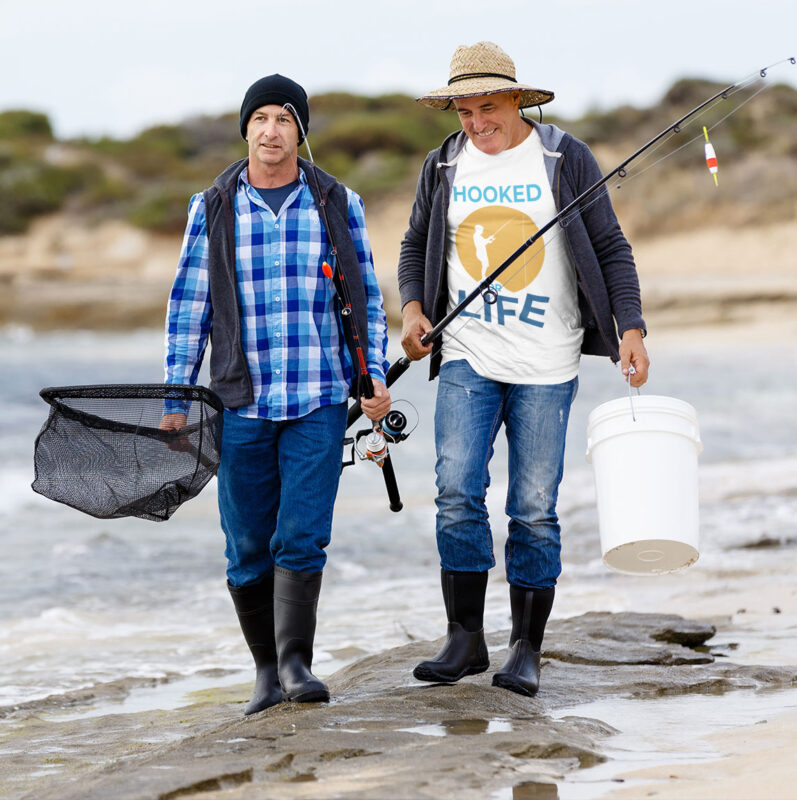 Two men walking along a beach with fishing gear. One man is wearing a plaid shirt, a vest, and black boots, carrying a fishing net. The other man is wearing a "Hooked for Life" fishing t-shirt, a straw hat, and black boots, carrying a white bucket and a fishing rod. Both are enjoying their time on the water.