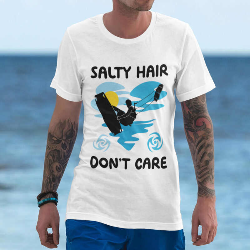 A person wearing a white 'Salty Hair, Don't Care' T-shirt with a silhouette of a kiteboarder in front of a beach and ocean. The shirt features the text 'Salty Hair, Don't Care' above a design of waves and a sun.
