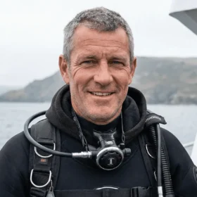 Smiling middle‑aged scuba diver in a black wetsuit on a boat, with the ocean and rocky coastline in the background.