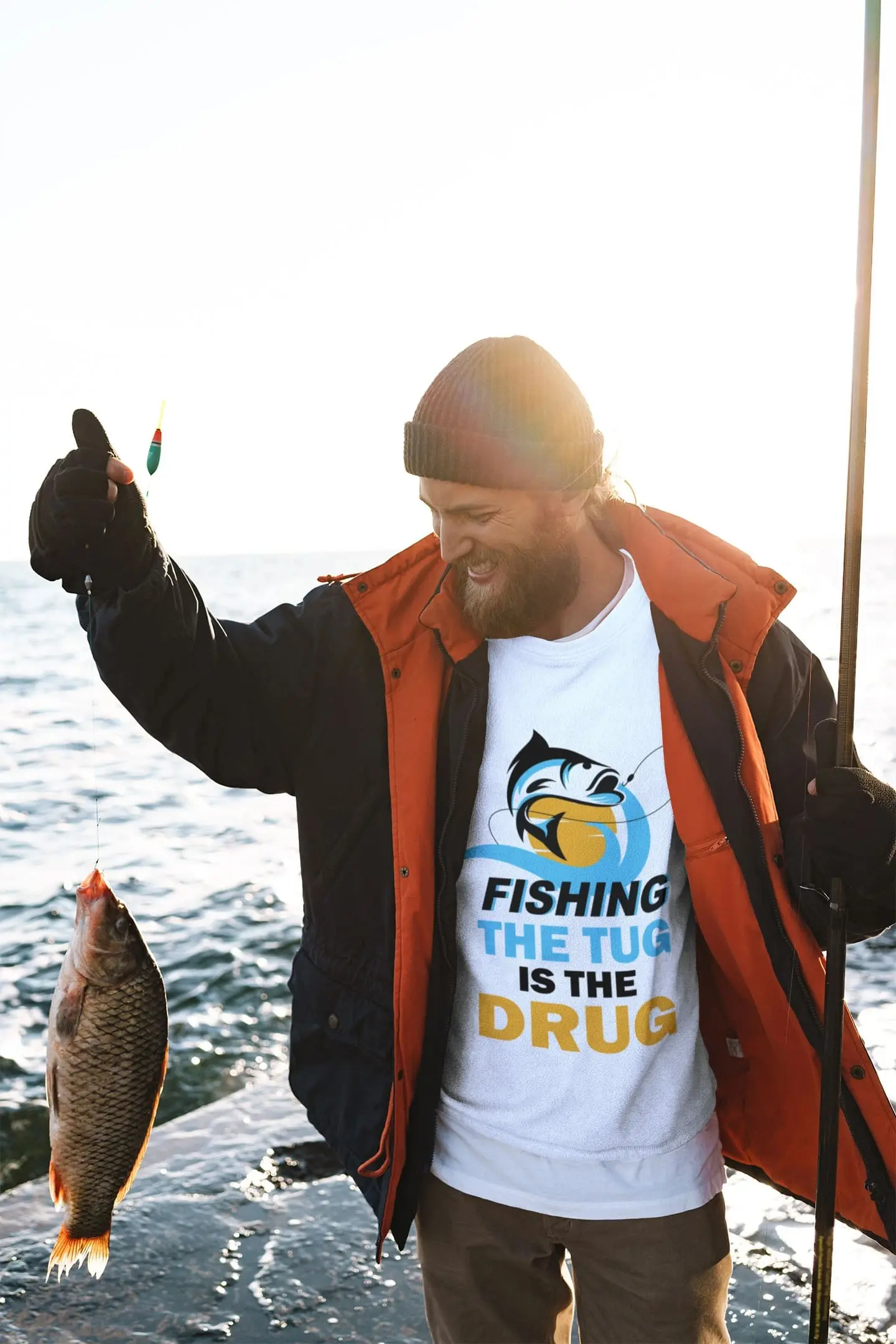 Man standing on a rocky shoreline holding a fish and wearing a white "Fishing the Tug Is the Drug" fishing t-shirt