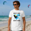 Kitesurfing Graphic Tee worn by a man standing on a sandy beach. The white shirt features a "Salty Hair Don't Care" graphic silhouette of a kiteboarder catching air against a blue heart-shaped wave and a yellow sun, with the ocean and kitesurfers visible in the background.