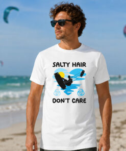 Kitesurfing Graphic Tee worn by a man standing on a sandy beach. The white shirt features a "Salty Hair Don't Care" graphic silhouette of a kiteboarder catching air against a blue heart-shaped wave and a yellow sun, with the ocean and kitesurfers visible in the background.