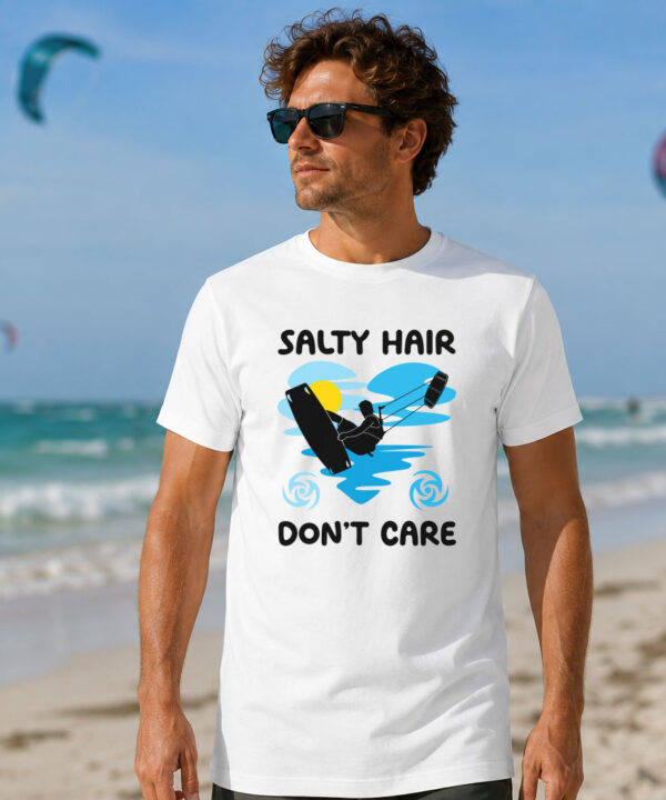Kitesurfing Graphic Tee worn by a man standing on a sandy beach. The white shirt features a "Salty Hair Don't Care" graphic silhouette of a kiteboarder catching air against a blue heart-shaped wave and a yellow sun, with the ocean and kitesurfers visible in the background.