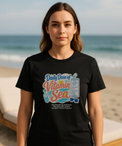 Woman standing on a sunny beach wearing a black premium beach graphic tee with a retro vitamin sea supplement print