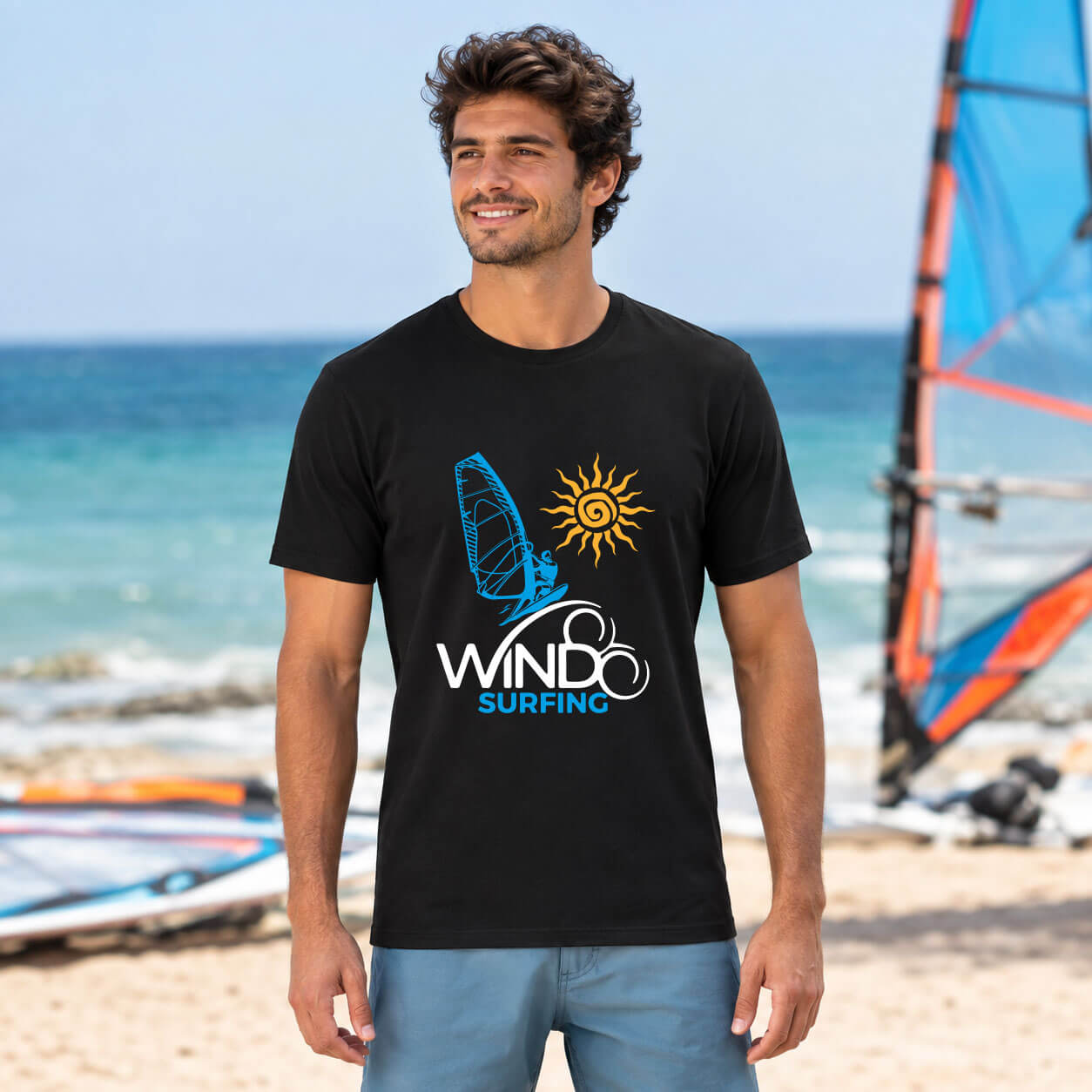 Man wearing the black Seasportees windsurfing t-shirt standing on a sunny beach with windsurfing gear in the background.